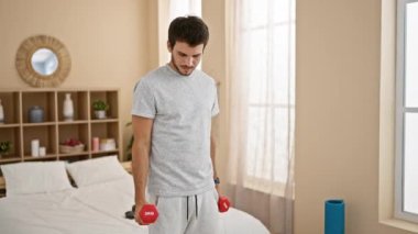 Odaklanmış bir adam, iyi aydınlatılmış bir yatak odasında kırmızı dambılları kaldırır, ev antrenmanını canlandırır.