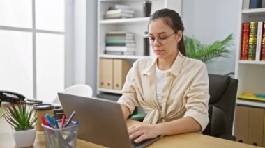 Kendine güveni tam genç İspanyol kadın çalışan, ofis masasında kollarını açıyor, dizüstü bilgisayarında yoğun bir şekilde çalışırken duraksıyor, zarif ofis içinde güzel bir gülümseme yayıyor.