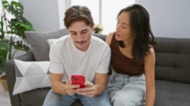 Irklar arası sevgi dolu bir çift, kadın ve erkek, evde rahat, gri bir kanepede, rahat bir oturma odasında, akıllı bir telefonla bir anı paylaşıyorlar..