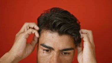 Genç bir İspanyol adam, saçlarını izole edilmiş kırmızı bir duvara dayar. Kişisel bakım ve tarzını yansıtır..