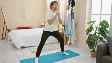 Açık renk bir yatak odasında sakallı genç bir adam mavi bir minder üzerinde yoga yapıyor. Evindeki huzuru ve huzuru arttırıyor..