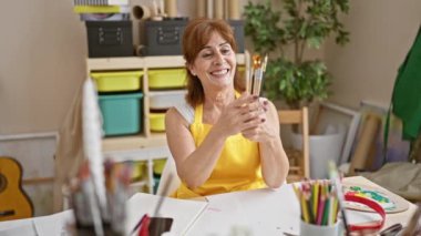 Heyecanlı, orta yaşlı kadın sanatçı zaferi gururlu bir gülümsemeyle kutluyor, elinde fırçalarla stüdyoda zafer için kollarını kaldırıyor.