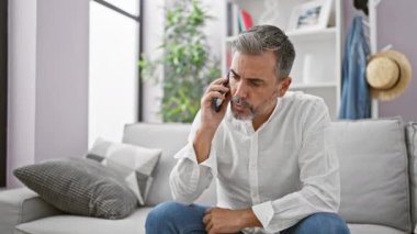 Genç İspanyol beyaz saçlı adam telefonda konuşuyor. Evdeki kanepede oturuyor.