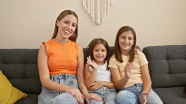 Gülümseyen genç bir kadın ve iki kız rahat bir şekilde gri bir koltukta rahat rahat oturma odasında oturmuş, bir aile anını resmediyorlar..