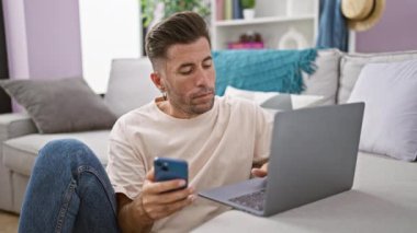 Çekici, İspanyol asıllı genç adam, modern online teknolojiye dalmış, rahat oturma odasında yerde otururken dizüstü bilgisayar ve akıllı telefon kullanmaya odaklanmış..