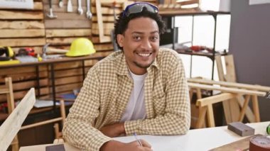 Gülümseyen genç adam iyi donanımlı bir marangozluk atölyesinde plan yapıyor.