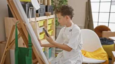 Sevimli sarışın sanatçı çocuk, akıllı telefona bakıyor, rahat resim atölyesinde resim yapmayı öğreniyor.