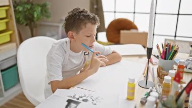 Yorgun küçük sarışın sanatçı çocuk, resim stüdyosunun masasına yaslanıyor, renkli resim sınıfının ortasında çocuk dostu öğrenme merkezinde sıkılıyor..