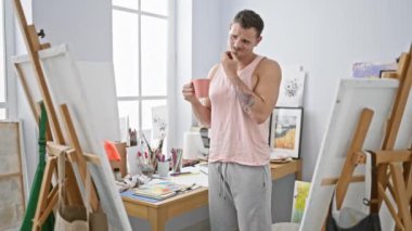 Dövmeleri olan düşünceli bir adam güneşli bir sanat stüdyosunda elinde bir kupayla oturuyor, sehpa ve yaratıcı malzemelerle çevrili..