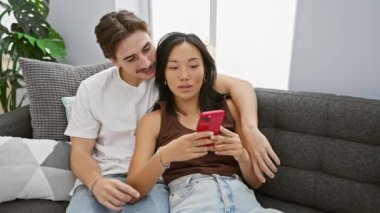 Irklar arası bir çift, bir erkek ve bir kadın, modern oturma odalarında birlikte akıllı telefon kullanarak bir neşe anlarını paylaşıyorlar..