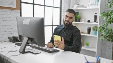 Modern ofis işyerinde akıllı telefon ve bilgisayar kullanan sakallı bir adam hayal kırıklığı ve konsantrasyon gösteriyor..