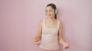 Spor kıyafetleri içinde, mavi gözlü ve kulaklıklı, izole edilmiş pembe arka planda şüpheli bir ifade gösteren cahil genç bir kadın..