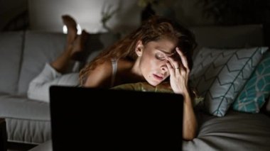 Geceleri oturma odasında dizüstü bilgisayar kullanan yorgun bir kadın evde stres ve yorgunluk gösteriyor.