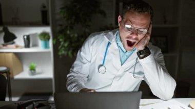 Gözlüklü ve laboratuvar önlüklü yorgun bir adam geç saatlere kadar klinikte dizüstü bilgisayarla çalışırken esniyor..