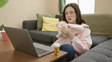 Pembe kazaklı beyaz bir kadın evinde laptopuyla ve ilaçlarıyla teletıp muayenesi yapıyor..