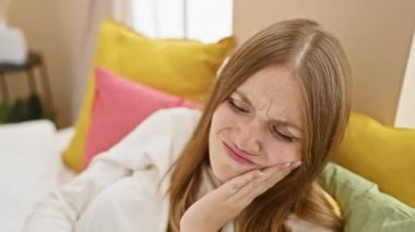 Diş ağrısı, dayanılmaz diş ağrısı çeken pijamalı genç sarışın bir kadın, elleri ve yüzü acı dolu bir şekilde yatakta oturuyor.