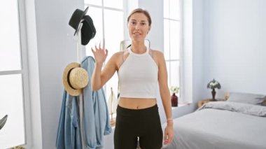 Askılı tişörtlü beyaz bir kadın, parlak, modern bir yatak odasında poz veriyor. Sağlık ve yaşam tarzını anlatıyor..