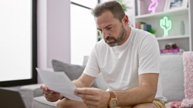 Beyaz tişörtlü, orta yaşlı yakışıklı bir adam modern oturma odasında gazete okuyor..