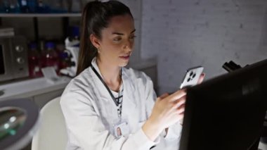 Laboratuvarda gülümseyen bilgisayar ve akıllı telefon kullanan güzel İspanyol bilim kadını.