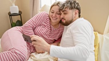 Mutlu bir çift, rahat bir yatak odasında yatarken birlikte akıllı telefona bakıp gülümsüyor.