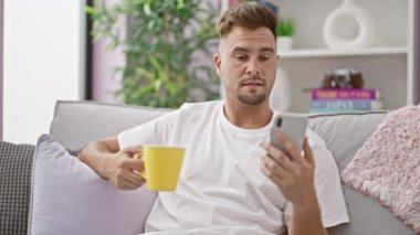 Akıllı telefona bakarken elinde kupa olan şok olmuş genç adam koltukta şaşkınlığını dile getiriyor..