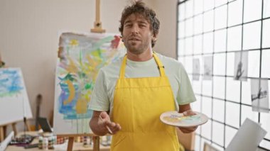 Sarı önlüklü düşünceli bir adam parlak bir sanat stüdyosunda resim yapıyor..