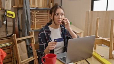 İspanyol kadın telefonda konuşuyor, elinde kredi kartı var. Marangozluk atölyesinde dizüstü bilgisayarı ve kupası var.
