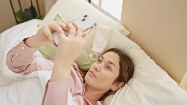 Rahat, sakin bir kadın yatakta uzanıyor, akıllı telefonunu parlak, rahat bir yatak odasında kullanıyor..