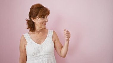 Neşeli orta yaşlı kadın neşeyle bir şeyler sunuyor. Elbise giyiyor ve avuç içiyle el kol hareketi yapıyor. Ayakta, gülümseyen, soyutlanmış pembe arkaplanı işaret eden sevimli görünümlü.