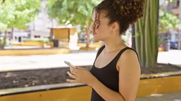 一位头发卷曲的年轻女子站在一片茂密的公园里用她的智能手机 — 图库视频影像