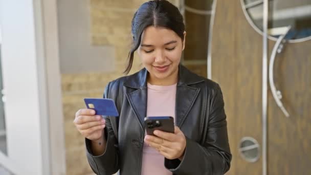 Hispanic woman checks credit card and smartphone in urban street setting.
