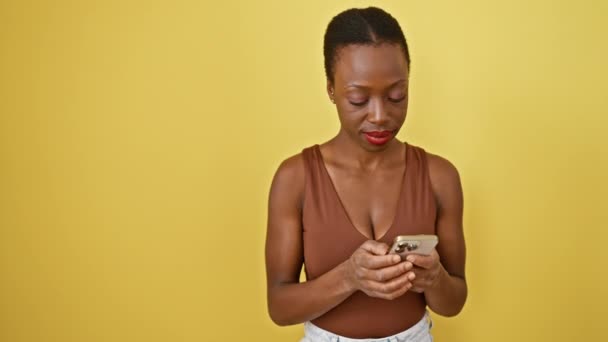 陽気なアフリカ系アメリカ人女性は 親指で側面を指し 口を開いた大きな笑顔 活気に満ちた隔離された黄色の背景に対して彼女のスマートフォンを使用して喜んで — ストック動画