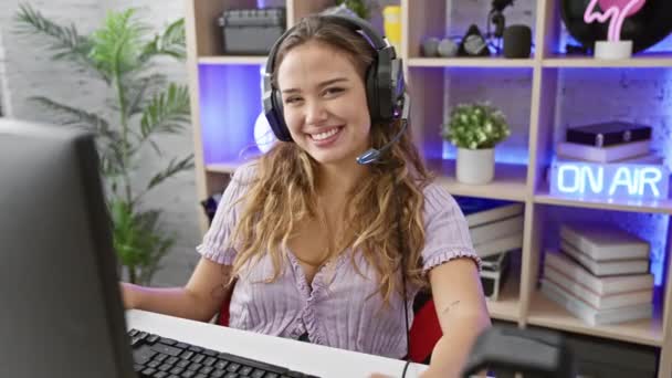 ジョイパックの若いヒスパニック女性ストリーマーは コンピュータ上でゲームをプレイし 口を開いた親指を与え 薄暗く点灯したゲームルームで耳から耳から耳まで微笑みます — ストック動画