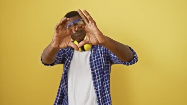 戴着安全眼镜的英俊的非洲裔美国男人站在孤立的黄色背景下 用象征性的心形手势示爱 在这个浪漫的概念中散发着幸福 — 图库视频影像