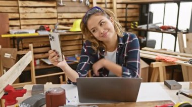 Enfes genç İspanyol kadın marangoz marangozluk stüdyosunda büyüleyici bir video görüşmesi yaptı, marangozluk becerilerini ve çekici saç stilini gösterirken gülümsüyordu.