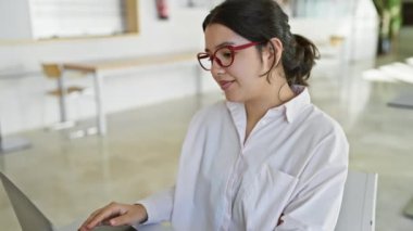 Genç İspanyol bir kadın, parlak, modern bir ofiste dizüstü bilgisayarıyla çalışıyor. Kırmızı gözlükleriyle sahneye renk katıyor..