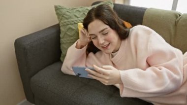 Pembe kazaklı neşeli genç bir kadın evdeki gri bir kanepede keyfi yerinde bir akıllı telefon kullanıyor.