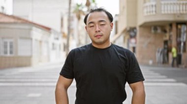 Neşeli genç Çinli adam, kendine güven ve neşe saçıyor, gülümsüyor ve hareketli bir şehir caddesinde çapraz kollar arasında dikilirken görülüyor.