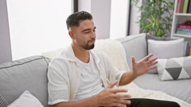 Öfkeli genç Arap adam, evde kanepede oturuyor, bağırıyor, çılgınca bir ifadeyle bağırıyor, kollarını kaldırıyor.