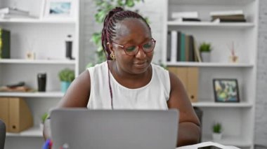 Kendine güvenen Afro-Amerikan iş kadını, dizüstü bilgisayar ve akıllı telefonla çalışıyor, ofisin içinde cesurca gülümsüyor.