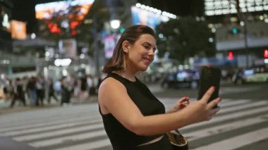 Gülümseyen İspanyol kadın, Tokyo 'nun parlak şehir sokaklarında güzel bir selfie çekiyor, şehir teknolojisi turizminin eğlencesini somutlaştırıyor..