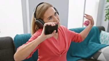Neşeli, olgun bir kadın, oturma odasında kulaklığı ve akıllı telefonuyla müzik dinlemekten hoşlanır..
