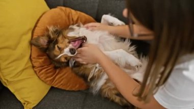 Genç bir İspanyol kadın, sevimli bir şekilde tüylü köpeğini renkli bir kanepede evcilleştiriyor, arkadaşlığını ve bakımını ön plana çıkarıyor..
