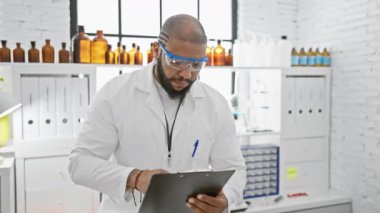 Modern bir laboratuvarda çalışan kendine güvenen siyah bilim adamı elinde bir dosya tutarken koruyucu gözlük takıyor.