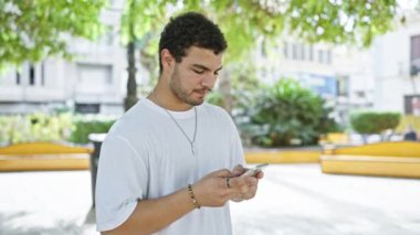 Düşünceli bir adam, güneşli bir parkta yemyeşil bir alanda akıllı telefonuyla iletişim kurar.