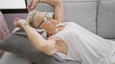 Gri saçlı, neşeli yaşlı kadın kanepede rahat bir şekilde uzanırken gülümsüyor. Mutlulukla ellerini başının üstüne koyuyor. Oturma odasındaki rahat güneş ışığının tadını çıkarıyor..