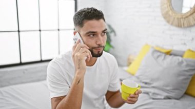 Beyaz tişörtlü, sakallı genç bir adam akıllı bir telefonla konuşuyor ve elinde parlak, modern bir yatak odasında kahve fincanı tutuyor..