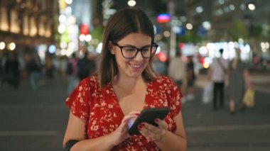 Neşeli ve güzel İspanyol bir kadın, neşeli bir anda yakalandı, akıllı telefonuyla şehir osaka sokağının gece ışıkları altında mesaj atıyor, bağlı bir dijital dünyanın özünü somutlaştırıyor..