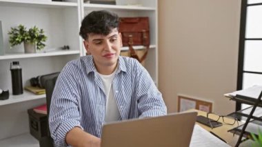 Modern ofis masasında gülümseyen genç adam dizüstü bilgisayarı, iç mekan bitkileri, kitaplar ve gözlükleriyle profesyonel ama sıradan bir atmosfer sağlıyor..