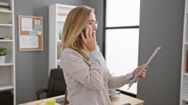 Gözlüklü bir iş kadını modern ofis ortamında telefonla konuşurken belgeleri inceliyor..